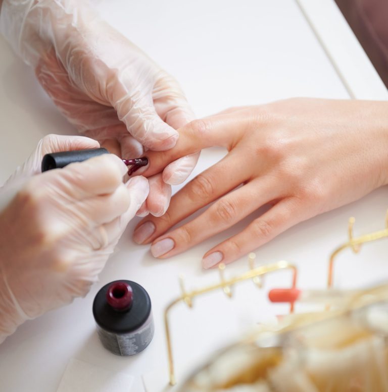 Professional manicurist doing manicure for woman client in beauty salon. Close up of manicure specialist hands in sterile gloves applying red nail polish on woman fingernail.